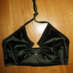 Mörkgrön sammetstopp med ringdetalj - Snygg mörkgrön croppad halterneck topp i mjuk sammet. Toppen har en cool ringdetalj framtill med rynkade partier och knyt runt nacken. Perfekt för dig som vill sticka ut med en unik och trendig look.