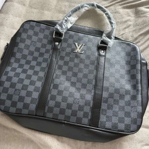 Datorväska i skinn - Snygg datorväska i svart och grått med klassiskt rutigt mönster. Väskan har silverfärgade detaljer, dubbla handtag och är tillverkad i skinn och syntet. Perfekt för att bära laptop och viktiga prylar med stil.