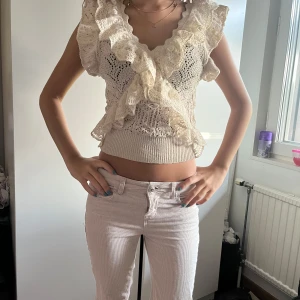 Beige spetsig volang blus - Supersöt beige blus med virkade detaljer och stora volanger framtill. Toppen har v-ringning och kan bäras både på axlarna och offshoulder. Den är croppad och har bred, ribbad mudd nedtill. Perfekt för dig som gillar romantisk och bohemisk stil.