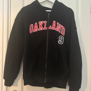 Svart Oakland hoodie med dragkedja - Svart hoodie med huva och dragkedja framtill. På bröstet står det 'OAKLAND' i rött och vitt samt siffran 9. Tröjan har känguruficka och långa ärmar. Perfekt för dig som gillar sportig och avslappnad stil.