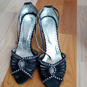 Svarta pumps med strass och öppen tå - Snygga svarta pumps med öppen tå och hög klack. Skorna har dekorativa strassdetaljer framtill och smala remmar som ger en elegant look. Materialet är satinliknande och insidan är silverfärgad. Perfekta för dig som vill sticka ut.De âr perfeckta att gå ut på ett party med.