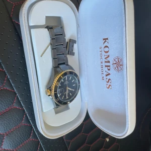 Svart och guldig klocka Kompass - Stilren klocka från Kompass Stockholm med svart urtavla, gulddetaljer och svart metallarmband. Klockan har tydliga index, datumvisning och vridbar bezel i guld och svart. Snyggt graverad baksida och lås med Kompass-logga. Perfekt accessoar för en modern look. Nypris 3000kr