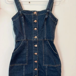 Jeansklänning med knappar från H&M Divided - Snygg jeansklänning från H&M Divided i mörkblå denim. Modellen är kort med breda axelband, knappar framtill och två stora fickor. Materialet är klassiskt jeans och passformen är normal. Säljer då den är för liten och aldrig använd.