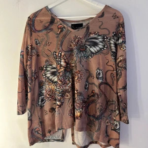 Mönstrad topp från KappAhl med fjärilar - Superfin topp från KappAhl i beige ton med unikt tryck av fjärilar, blommor och ormar. Trekvartsärm och v-ringad halsringning. Materialet är mjukt och stretchigt i polyester och elastan, perfekt för dig som gillar coola prints och bekväma plagg. Storlek M