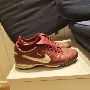 Röda Nike fotbollsskor Ronaldinho - Snygga fotbollsskor från Nike i rött skinn med guldiga detaljer och Ronaldinho-signatur. Skorna har klassisk snörning, låg profil och vit sula med svart yttersula. Var från början gjorda för street fotboll men är sjukt snygga som bara sneakers. otroligt bra skick för sin ålder och använda som golf skor innan så under sulan är som ny.