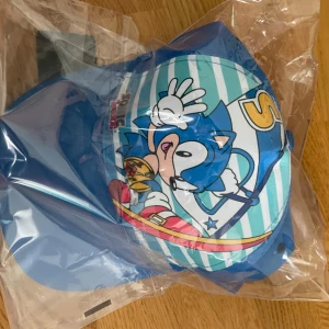 Blå Sonic the Hedgehog keps - Helt nya Sonic the Hedgehog keps i klarblå färg från Sun City Sonic. Framsidan har ett stort tryck med Sonic och vita ränder i bakgrunden. Kepsen har böjd skärm och justerbar passform. Perfekt för dig som älskar gaming och coola accessoarer.