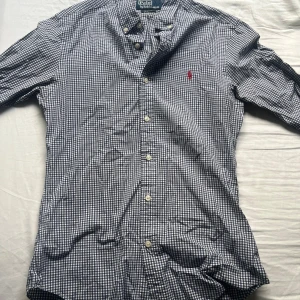 Rutig skjorta från Polo Ralph Lauren - Klassisk rutig skjorta från Polo Ralph Lauren i blått och vitt. Skjortan har lång ärm, knappar hela vägen och den ikoniska röda loggan broderad på bröstet.