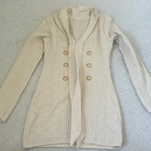 Beige klänning med guldknappar och knyt - Snygg beige klänning med strukturerat mönster och sex dekorativa guldknappar framtill. Koftan har lång ärm, v-ringning och ett elegant knytband vid halsen. Perfekt för dig som gillar en chic och stilren look.