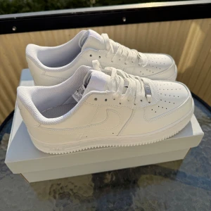 Nike Air Force 1 helvita sneakers - Klassiska Nike Air Force 1 i helvitt skinn med perforerad tå, vit sula och snörning. Stilren och tidlös modell som passar till allt. Snygg låg siluett och ikonisk Nike-logga på sidan. Perfekta för dig som gillar cleana sneakers med streetkänsla.