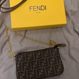 Fendi brun axelväska med FF-mönster - Säljer en brun axelväska från Fendi med ikoniskt FF-mönster i svart. Väskan har guldfärgad kedja, dragkedja och detaljer i skinn. Perfekt storlek för det viktigaste och har en snygg, lyxig vibe.