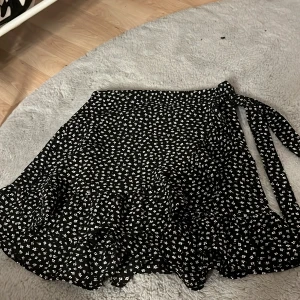 Svart blommig omlottkjol FB Sister XS - Supersöt svart kort omlottkjol från FB Sister i storlek XS. Kjolen har små vita blommor, volangkant och knytband i midjan. Perfekt för dig som gillar en feminin och trendig stil med extra detaljer.