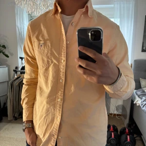 Orange oxfordskjorta från Gant M - Säljer en klassisk oxfordskjorta från Gant i orange bomull. Skjortan har button-down krage, bröstficka med broderad Gant-logga och vita knappar. Passformen är normal och ärmarna är långa. Perfekt för en clean och stilren look.