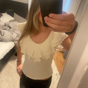Vit blus med volang och v-ringning - Supersöt vit blus med v-ringning och stora volanger över axlar och bröst. Toppen är smockad och sitter tajt över kroppen, vilket ger en snygg siluett. Perfekt för dig som gillar romantiska detaljer och en feminin look.