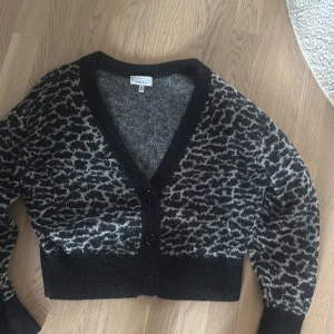 Grå kofta med leopardmönster - Snygg grå och svart kofta från American Vintage med leopardmönster. Croppad modell med v-ringning och tre svarta knappar framtill. Mjuk och stickad känsla, perfekt för lager på lager. Långärmad och passar till många olika stilar.
