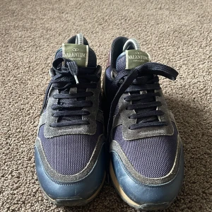 Valentino Rockrunner sneakers blå/grå - Snygga Valentino Rockrunner sneakers i marinblått mesh, grå mocka och blått skinn. Sulan har ikoniska nitar och baksidan är i vinrött skinn. Skorna har snörning och coola kontraster mellan material och färger. Perfekta för dig som gillar lyxiga och unika sneakers.tyvär är allt annat slängt men skorna är ett super bra sick med några effekter. Bara skorna till kommer vid köp. Mer frågor kom och prata försöker vara så snabb så möjligt 