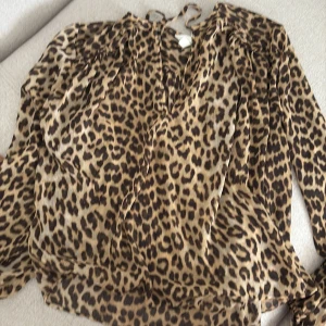 Leopardmönstrad blus från H&M - Säljer en luftig blus från H&M med leopardmönster i bruna och svarta toner. Blusen har långa ärmar, v-ringning med knytband i halsen och är tillverkad i ett tunt, flowy material som känns riktigt skönt mot huden.