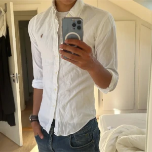 Ralph Lauren Skjorta (pris diskuterbart) - !! NY SKICK !! Klassisk vit skjorta från Ralph Lauren med button-down krage i SLIMFIT och broderad logga på bröstet. Skjortan har lång ärm, tillverkad i bomull. Perfekt till jeans för en clean och stilren look. storlek 20 Sitter som en S-M