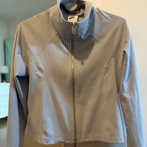 Ljusblå vindjacka från Nike - Snygg ljusblå vindjacka från Nike med hel dragkedja framtill och hög krage. Jackan har en clean och sportig look, perfekt för dig som gillar stilrena plagg. Tillverkad i mjukt och lätt material som passar för aktiva dagar.
