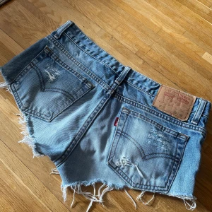 Levi's ljusblå slitna jeansshorts - Säljer ett par ljusblå jeansshorts från Levi's med råa, fransiga kanter och slitningar både fram och bak. Klassisk femficksmodell med knapp och dragkedja. Perfekta för sommaren och ger en cool, avslappnad vibe. Står inte storlek men som S/M