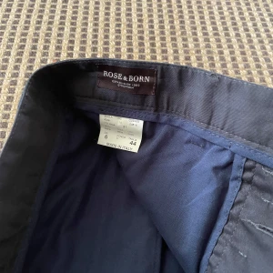 Chinos från Rose & Born - Snygga svarta kostymbyxor från Rose & Born, tillverkade i Italien. Klassisk rak modell med diskreta fickor och bälteshällor. Perfekta för en clean och stilren look. Materialet känns mjukt och har en lätt glans.