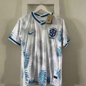 Vit Brasilien fotbollströja Nike M - Snygg Brasilien fotbollströja från Nike i vitt med blå och grå palmbladsmönster. Tröjan har korta ärmar, blå detaljer vid krage och ärmslut samt broderat CBF-märke och Nike-logga på bröstet. Tillverkad i lätt och ventilerande polyester.