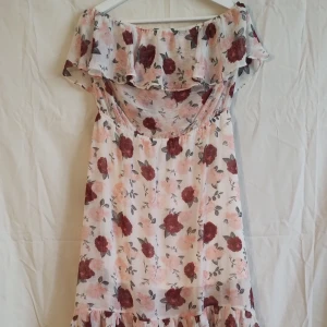 Blommig offshoulder klänning H&M Divided - Söt midiklänning från H&M Divided med offshoulder och volangdetaljer. Klänningen är vit med blommigt mönster i vinrött och ljusrosa, tillverkad i ett tunt, luftigt polyestertyg. Perfekt för sommarens alla tillfällen!