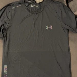 Svart tränings-tshirt från Under Armour - Svart kortärmad t-shirt från Under Armour med diskret logga på bröstet och textdetaljer på axeln. Tillverkad i lätt och teknisk polyester, perfekt för träning. Snygg passform och rund halsringning.