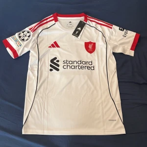Liverpool bortatröja Virgil 4 Adidas - Liverpool FC bortatröja från Adidas med Virgil van Dijk #4 på ryggen. Vit med röda detaljer, svarta linjer och klubbmärke på bröstet. Kortärmad, tillverkad i lätt funktionsmaterial. UEFA Foundation-märke på ärmen och Standard Chartered som sponsor framtill.