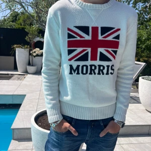 Vit stickad tröja Morris Union Jack - Vit stickad tröja från Morris med Union Jack-flagga och tydlig logga framtill. Tröjan har ribbade muddar vid ärmslut och nederkant samt rund halsringning. Perfekt för dig som gillar klassisk brittisk stil med en modern twist.