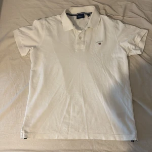 Vit pikétröja från GANT, storlek S - Klassisk vit pikétröja från GANT med korta ärmar och krage. Tröjan har broderad GANT-logga på bröstet och knappar vid halsen. Tillverkad i mjuk bomull, perfekt för en clean och stilren look.