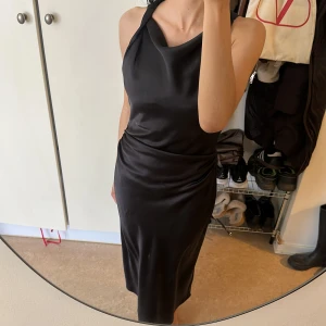 Svart satinklänning med drapering - Supersnygg svart satinklänning med draperad halsringning och smickrande rynkdetalj vid midjan. Klänningen är ärmlös och har en elegant, figurnära passform som ger en stilren siluett. Perfekt för dig som vill ha en chic och tidlös look. Några slitningar, fråga för fler bilder❤️❤️