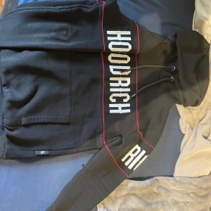 Svart HOODRICH hoodie med röda detaljer - Svart hoodie från HOODRICH med stora vita tryck på bröstet och ärmen. Snygga röda linjer längs överdelen och ärmarna. Klassisk huva med snörning och stor magficka framtill. Använt bara 3-6 gånger blev för liten för mig väldigt snabbt.