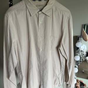 Beige skjorta från Burberry Brit - Stilren beige skjorta från Burberry Brit i 100% bomull. Klassisk krage, bröstficka och knäppning framtill. Snygga rutiga detaljer på insidan av kragen och ärmsluten ger en extra touch. Perfekt för dig som gillar en clean och tidlös look.