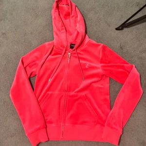 Juicy Couture rosa velour hoodie - Juicy Couture hoodie i mjuk rosa velour med dragkedja framtill, snörning i huvan och klassisk JC-logga broderad på bröstet. Känguruficka och ribbade muddar ger en skön och trendig look. Perfekt för dig som gillar comfy och stilrent.