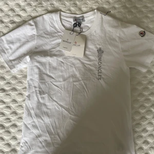 Vit Moncler t-shirt med broderad logga - Vit t-shirt från Moncler i mjuk bomull med broderad Moncler-logga i grått på bröstet och klassisk patch på ärmen. T-shirten har rund halsringning och korta ärmar. Perfekt för dig som gillar stilrena och exklusiva plagg.