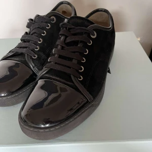 Lanvins - Snygga svarta sneakers från Lanvin med lyxig känsla. Skorna har en glansig lackad tå och ovandel i mjuk mocka, samt svarta snören och metallöljetter. Perfekta för dig som gillar stilrena och exklusiva detaljer.