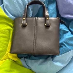 Svart handväska med gulddetaljer - Stilren svart handväska i skinnimitation med två handtag och snygga guldfärgade detaljer. Väskan har dragkedja upptill och flera fack inuti för att hålla ordning på dina grejer. Perfekt storlek för att bära med sig det viktigaste.