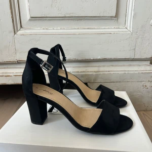 Svarta klackade sandaler från Alley - Stilrena svarta sandaler från Alley med bred rem över tårna och justerbart spänne runt ankeln. Skorna har en hög blockklack och öppen tå, tillverkade i mockaliknande material. Perfekta för att lyfta din outfit med en elegant touch.