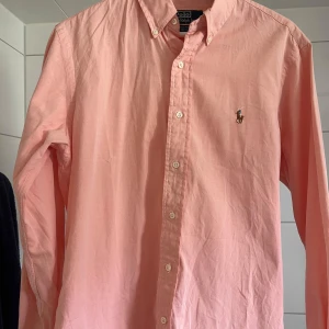 Rosa skjorta från Polo Ralph Lauren - Storlek L men ganska liten i storleken. Snygg rosa skjorta från Polo Ralph Lauren med klassisk button down-krage och broderad logga på bröstet. Skjortan har lång ärm, custom fit och stängs med vita knappar framtill. Perfekt för sommaren.