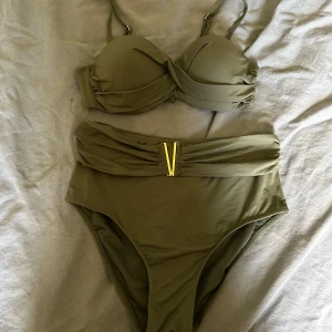 Olivgrön bikini med hög midja och gulddetalj - Snygg olivgrön bikini med vadderad bygel-bh och justerbara axelband. Bikinitrosan är high waist med en guldfärgad V-detalj framtill. Båda delarna har en stilren och enkel design, perfekt för strandhäng. Materialet är mjukt och stretchigt syntet. 