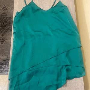 Turkos linnetopp från MNG Suit Collection - Säljer en turkos linnetopp från MNG Suit Collection i storlek XS. Toppen har smala axelband, v-ringning och dubbla lager tyg med asymmetrisk volangdetalj framtill. Materialet är glansigt och känns lyxigt, perfekt för att sticka ut med en färgklick.