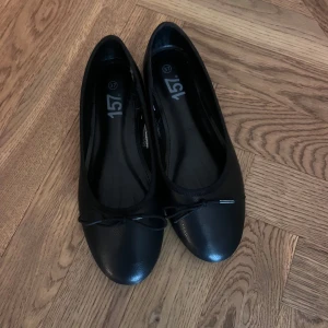 Svarta ballerinaskor med rosett - Klassiska svarta ballerinaskor från 157 med rund tå och liten rosett framtill. Skorna har platt sula och är tillverkade i syntetmaterial med en stilren look som passar till det mesta. Perfekta för dig som gillar enkel och tidlös stil. Helt slutsålda överallt 