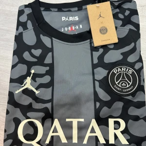 PSG x Jordan svart/grå fotbollströja - Snygg PSG x Jordan fotbollströja i svart och grått med unikt camouflageinspirerat mönster. Tröjan har korta ärmar, rund hals och tryckta loggor från både Jordan och PSG samt Qatar Airways som sponsor. Tillverkad i lätt och ventilerande material, perfekt för match eller träning.