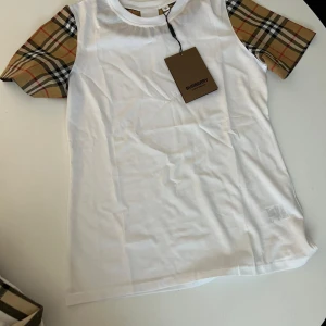 Vit Burberry t-shirt med rutiga ärmar - Snygg vit t-shirt från Burberry med klassiska rutiga korta ärmar i beige, svart och rött. T-shirten har rund halsringning och är gjord i mjuk bomull. Perfekt för dig som vill ha en stilren look med en touch av lyx.