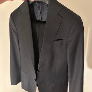 Svart kostym från Oscar Jacobson - Snygg svart slim kostym från Oscar Jacobson i klassisk modell. Kavajen är i strl 44 och byxorna i strl 44, perfekt för dig som är runt 170-175 cm. Skicket är väldigt bra och hör av dig om du vill ha mer specifika mått eller fler bilder.