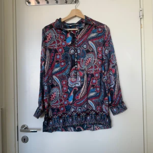 Färgglad paisleymönstrad henley - Paisleymönstrad henley-blus i blått, rött och vitt med långa ärmar och knappar framtill. Blusen har en lös passform och är tillverkad i ett lätt, mjukt material som känns skönt mot huden. Perfekt för dig som gillar färg och mönster.Från märket Odd Molly i storlek 1 som ungefär motsvarar en storlek S. Mycket fint skick, jag hittar inga synlig anmärkningar eller defekter. 