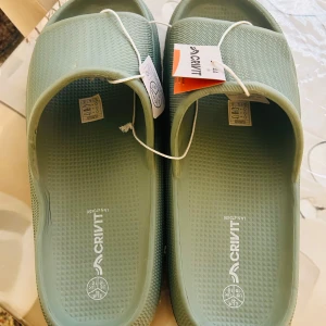 Gröna tofflor från Crivit stl 44 - Snygga ljusgröna sandaler/ tofflor från Crivit i storlek 44. Tillverkade i syntetmaterial med räfflad sula och öppen tå. Perfekta att ha ute eller i stranden, med enkel slip-on design och bekväm passform.