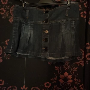 Mörk jeanskjol med knappar från Madonna - Snygg mörkblå lågmidjad mini jeanskjol från Madonna med stora knappar framtill och coola sömdetaljer. Kjolen är kort och har lätt utställd form med veck för extra volym. Perfekt för dig som gillar en trendig och avslappnad stil.