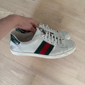 Gucci Ace vita sneakers med röd/grön rand - Gucci Ace sneakers i vitt skinn med klassiska gröna och röda ränder på sidorna. Baksidan har mörkgrön detalj med reptilstruktur. Platt vit sula och snörning framtill. Perfekta för dig som vill ha en clean men lyxig look. Dm för frågor🙌🤝