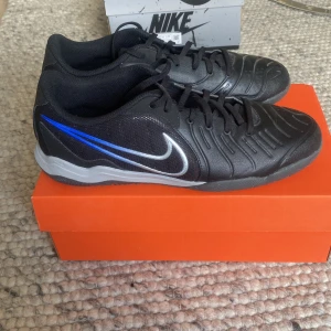 Nike Legend 10 Academy IC fotbollsskor - Nike Legend 10 Academy IC är svarta inomhusfotbollsskor med krom- och blå detaljer. Skorna har snörning, rund tå och platt sula. Ovandelen är i syntetmaterial med en cool blå Swoosh längs sidan. Perfekta för dig som vill sticka ut på planen.